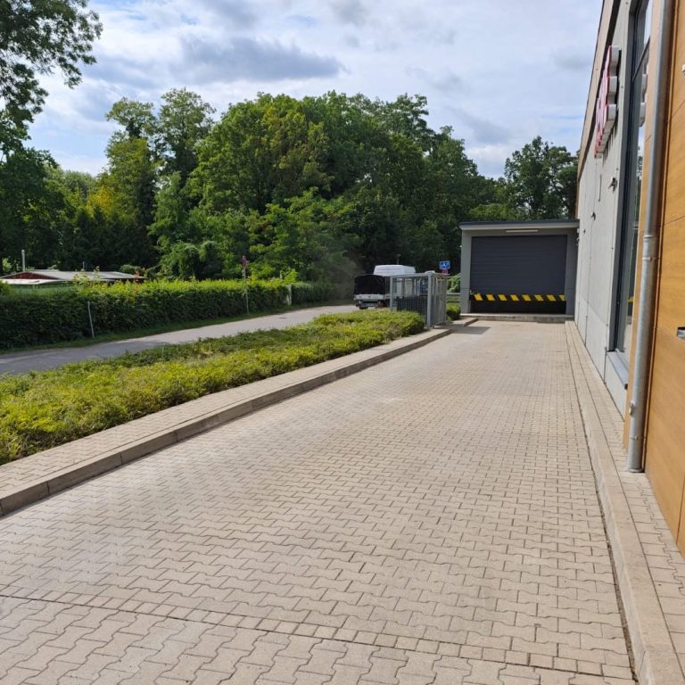 Grünpflege Zufahrtsbereich mit gepflastertem Weg und Tor, umgeben von grüner Vegetation.