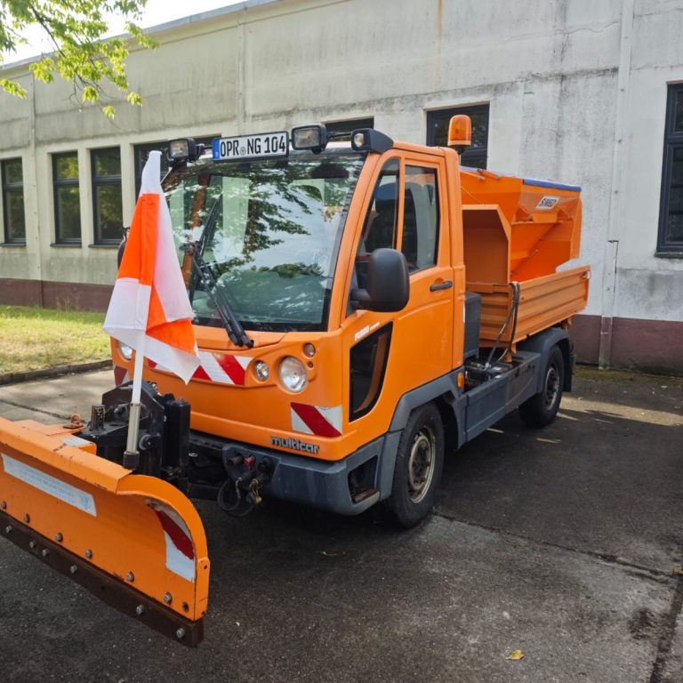 Multicar Orange-farbener Kommunalfahrzeug mit Schneepflug und Warnflagge.